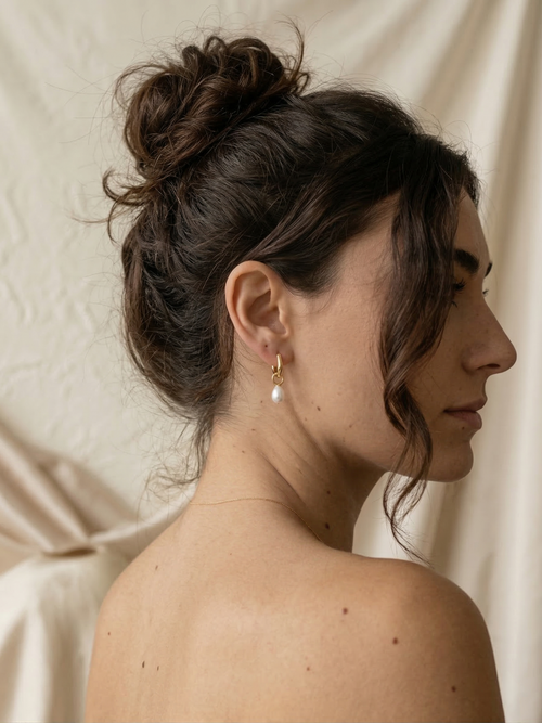 Close-up of a person wearing a gold earring with a pearl on a neutral background